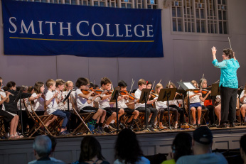 Student orchestra performs on stage facing the conductor in a bright blue blouse, while an audience in the foreground looks on.