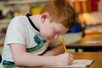 A boy in a t-shirt with a pencil in his hand and a determined expression is hunched over a paper. 