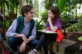 In the arboretum, a young man squats in front of a young girl sitting on a bench with a clipboard in her lap. The girl has her chin resting on her hand and appears to be thinking about something that she and the man are discussing,