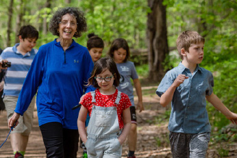 A group of children walking along a wide pathway in a wooded setting with a smiling teacher.