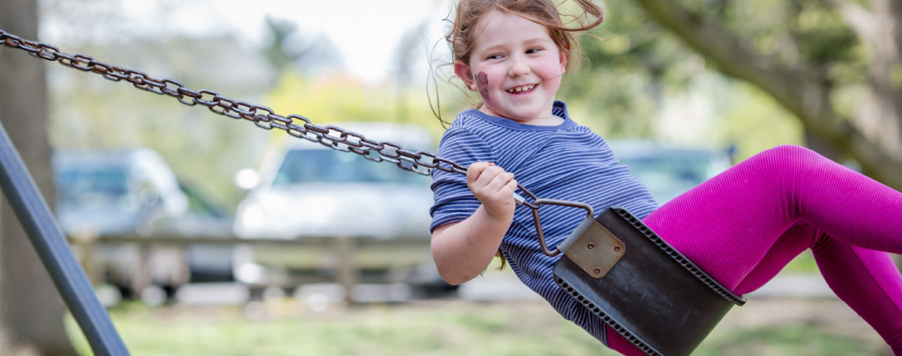 A girl on a swing wearing a blue t-shirt and bright pink tights and a smudge of dirt on her cheek, smiling as she looks to her right towards the camera. 