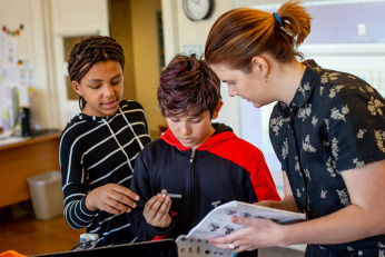 A boy is holding an object in his hand, as a girl behind him points at it and a woman facing them holds a book.