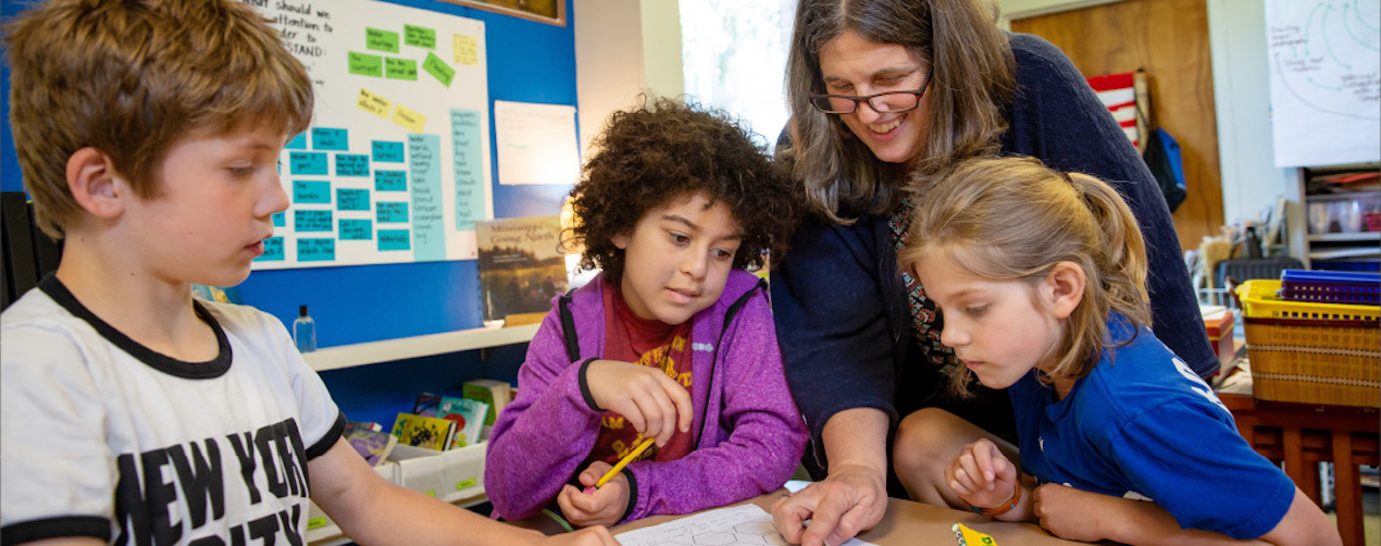 Two children are working together at a table as a teacher looks on.