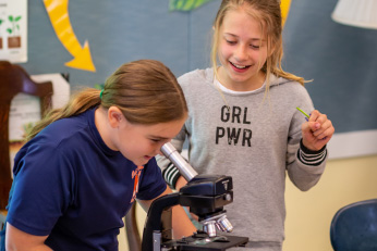 A girl looks into a microscope while a second girl watches with excitement.