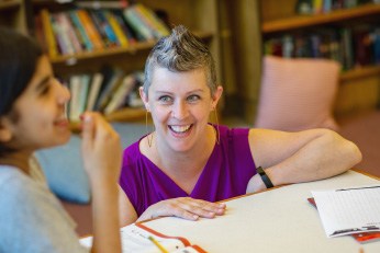 A woman is seated or kneeling behind a low table with a big smile as she gazes upward at a young girl in the left foreground.