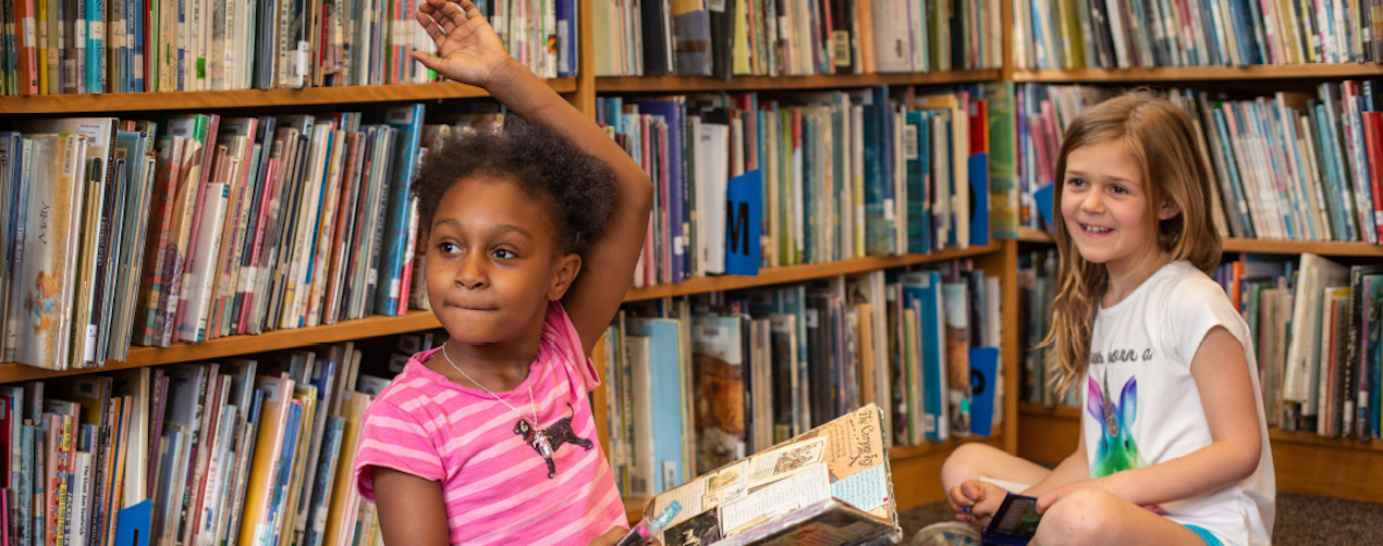 Two girls holding books are seated on the floor in front of a wall of bookshelves, looking at someone outside of the image. One girl has her hand raised.
