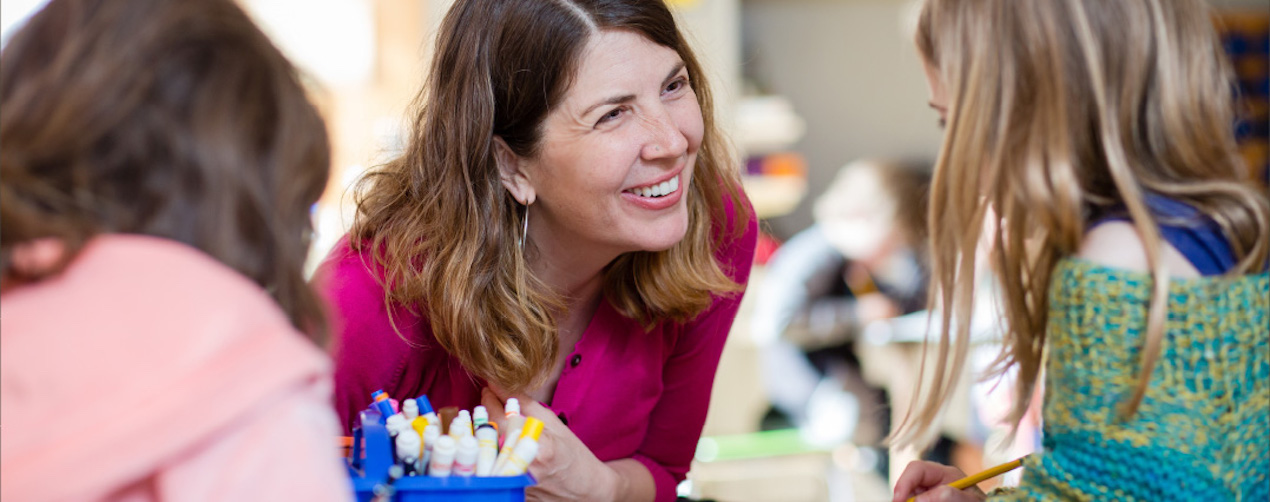 A female teacher facing the viewer smiles at two young girls with their backs to the viewer.