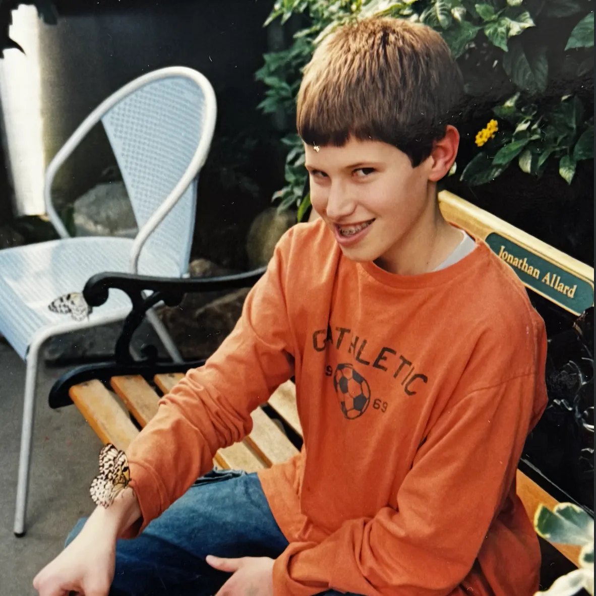 A young boy in an orange shirt smiles at the camera with a butterfly perched on his wrist